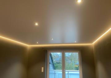 Spanndecke in einem Zimmer mit Fenster, umlaufender indirekter Beleuchtung und mehreren Einbauspots.